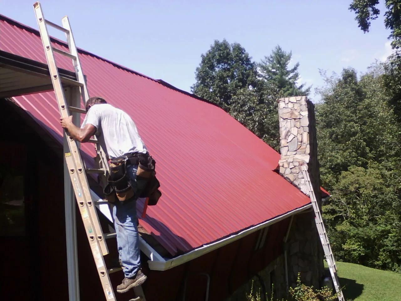Roofer on a ladder beside a red metal roof and stone chimney — Soffit & Fascia Repair by Roofing Brothers in Villanova